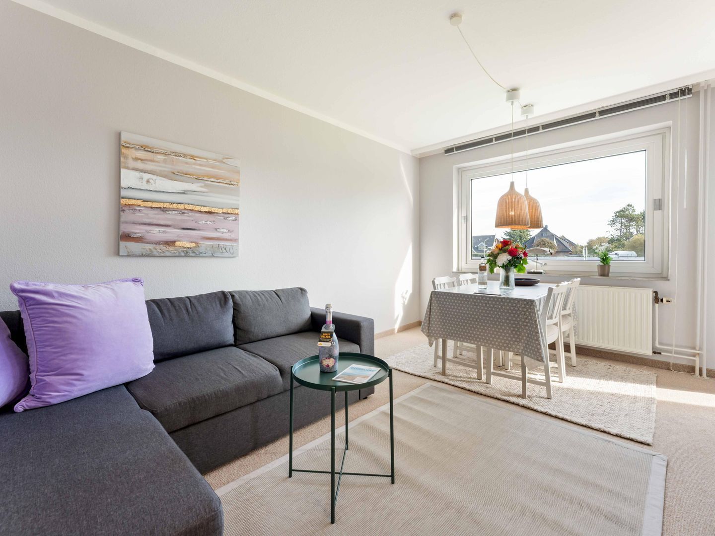 A modern living room with a grey sofa, a small round table with a bottle, and a dining area with a white table and chairs.