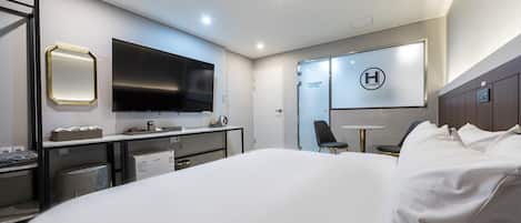 A modern hotel room with a large bed, a desk with a flat-screen TV, a seating area, and a minibar.