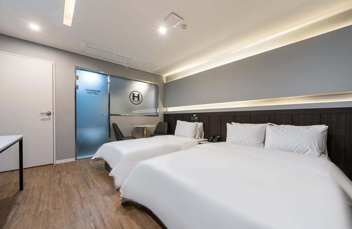 Een hotelkamer met twee bedden, een bureau, een stoel en een deur met de tekst 'Harbor Hotel'.