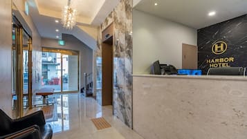Zona de recepción de un hotel con pared de mármol, mostrador de recepción y una araña.
