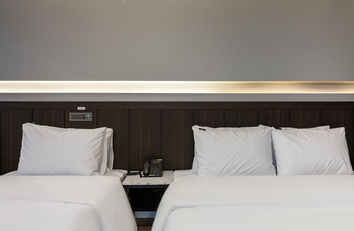 Habitación de hotel con dos camas, una mesita de noche con teléfono y un panel de control en la pared.
