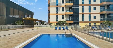 A swimming pool with a tiled deck, surrounded by lounge chairs and a high-rise building with balconies.