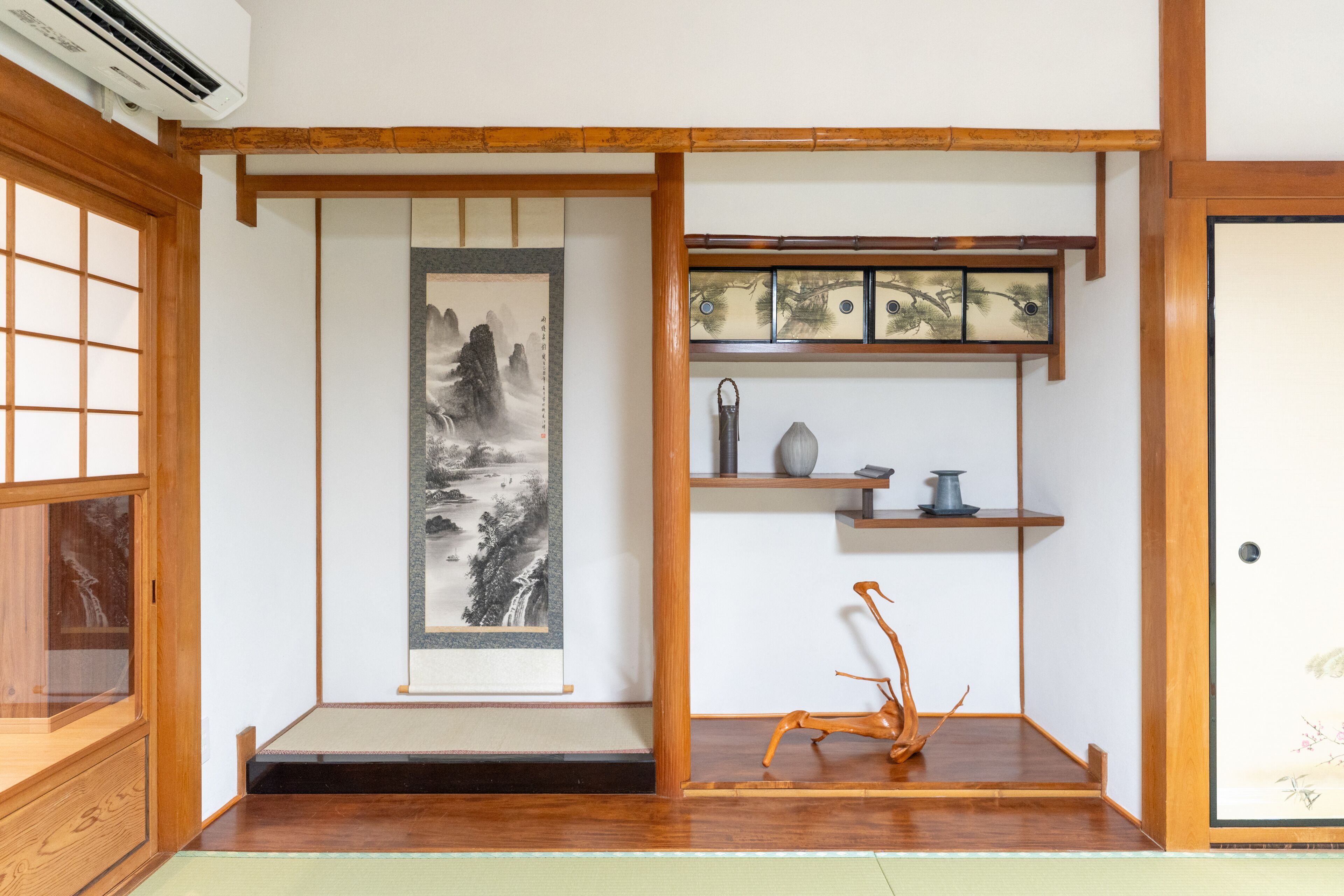 Una habitación japonesa tradicional con suelo de tatami, un panel shoji, un pergamino colgado, un estante pequeño con adornos y una puerta corredera.