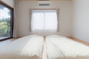 A room with two beds, a window with curtains, an air conditioning unit, and a view of greenery outside.