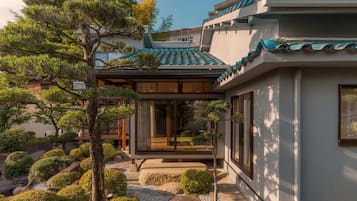 A traditional Japanese-style building with a tiled roof, surrounded by well-maintained landscaping including rocks, shrubs, and a pine tree.