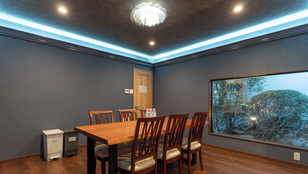 A dining area with a wooden table and chairs, a trash bin, and a window with a view of trees.