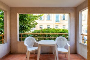 Ein Balkon mit zwei weißen Plastikstühlen und einem kleinen Tisch, mit Blick auf ein Gebäude mit einer grünen Hecke und blauem Himmel.
