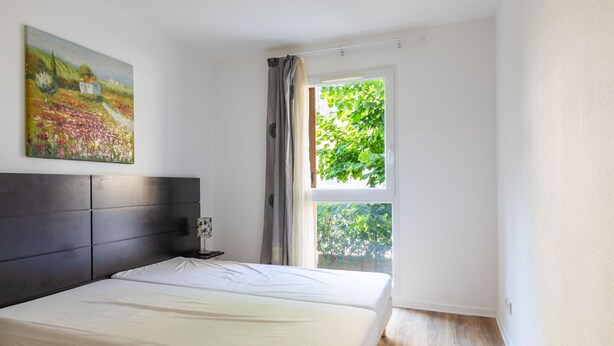 Un dormitorio con cama, mesita de noche, lámpara, un cuadro y ventana con vista a follaje.