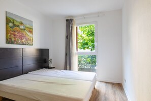 A bedroom with a bed, a nightstand, a lamp, a painting, and a window with a view of greenery. - Apartment with balcony and swimming pool, sleeps 4 (Grasse)
