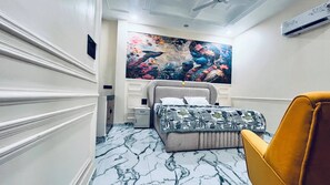 A hotel room with a marble floor, a bed with a patterned bedspread, a large floral painting on the wall, and a yellow armchair. - Mannat Villa (Ramnagar)