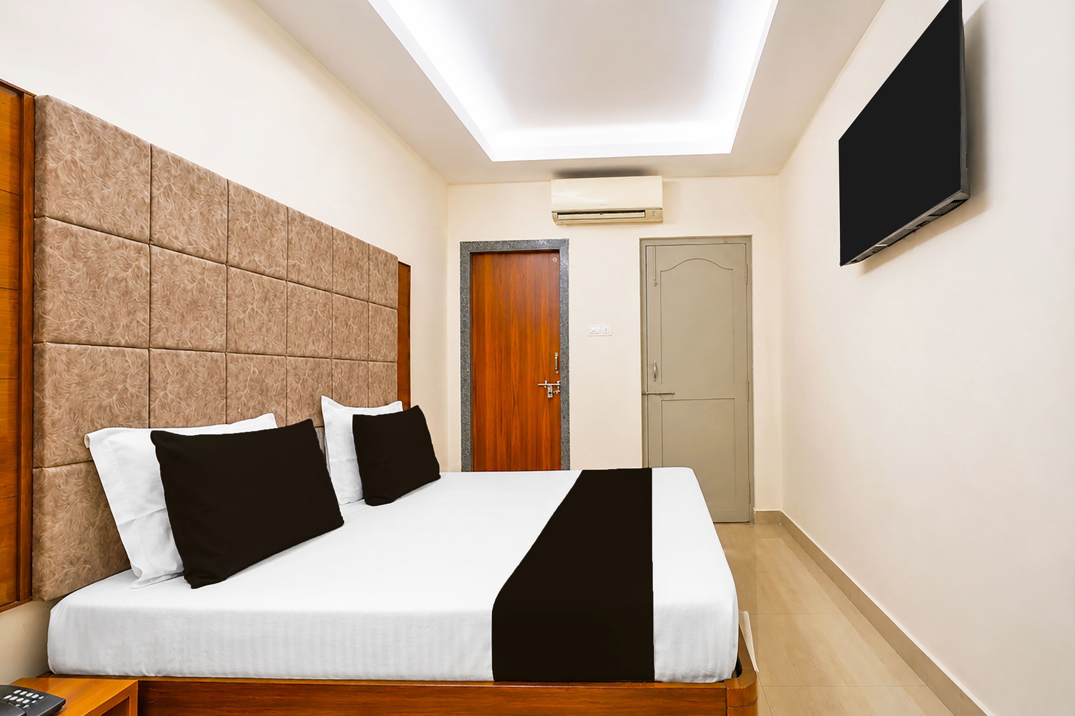 A hotel room with a bed, a flat-screen TV, an air conditioning unit, and a door.