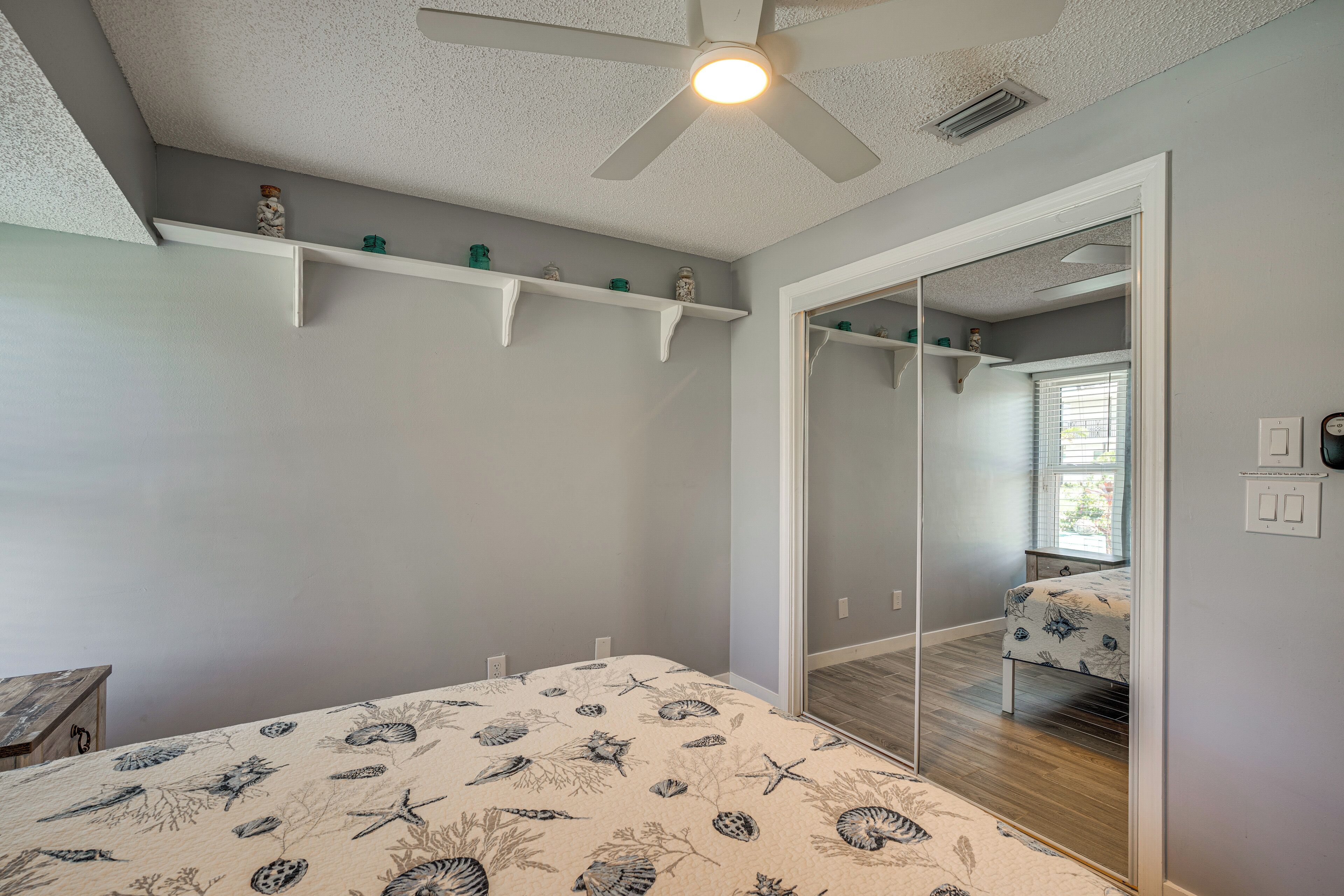 Une chambre à coucher avec un lit, un ventilateur de plafond, un miroir et une fenêtre avec des stores.