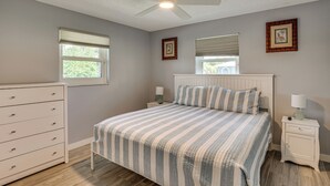 Une chambre à coucher comprenant un lit, une commode, une table de chevet, un ventilateur de plafond et deux fenêtres équipées de stores.