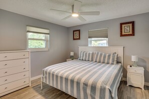 Ein Schlafzimmer mit einem Bett, einer Kommode, einem Nachttisch, einem Deckenventilator und zwei Fenstern mit Jalousien.