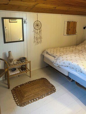 Quarto com cama, mesa de cabeceira com vaso e livros, um sonho apanhador, espelho e um armário de madeira.