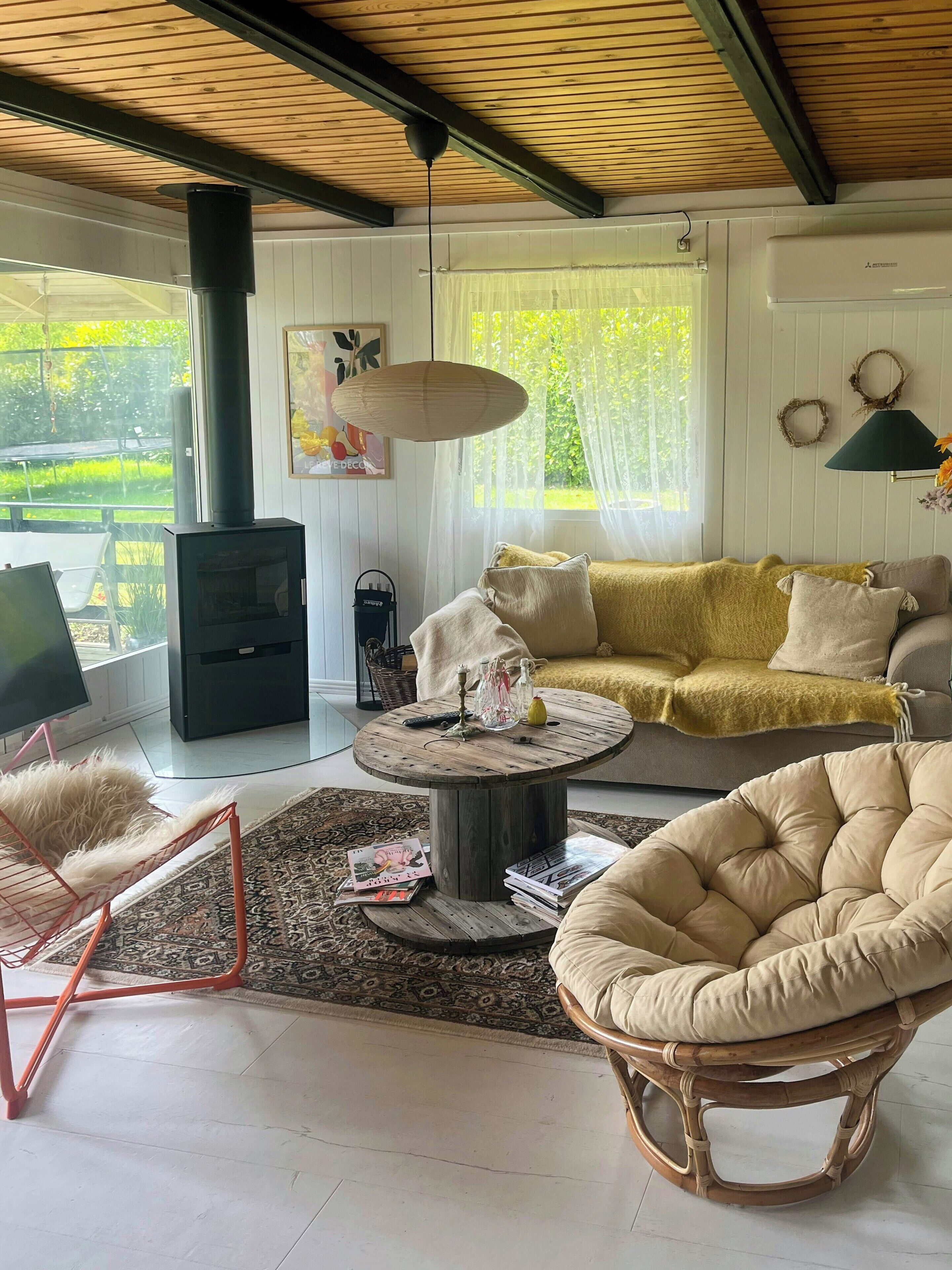 Ein gemütliches Wohnzimmer mit einer Couch, einem runden Couchtisch, einem Korbstuhl, einem Kamin und Blick ins Grüne.