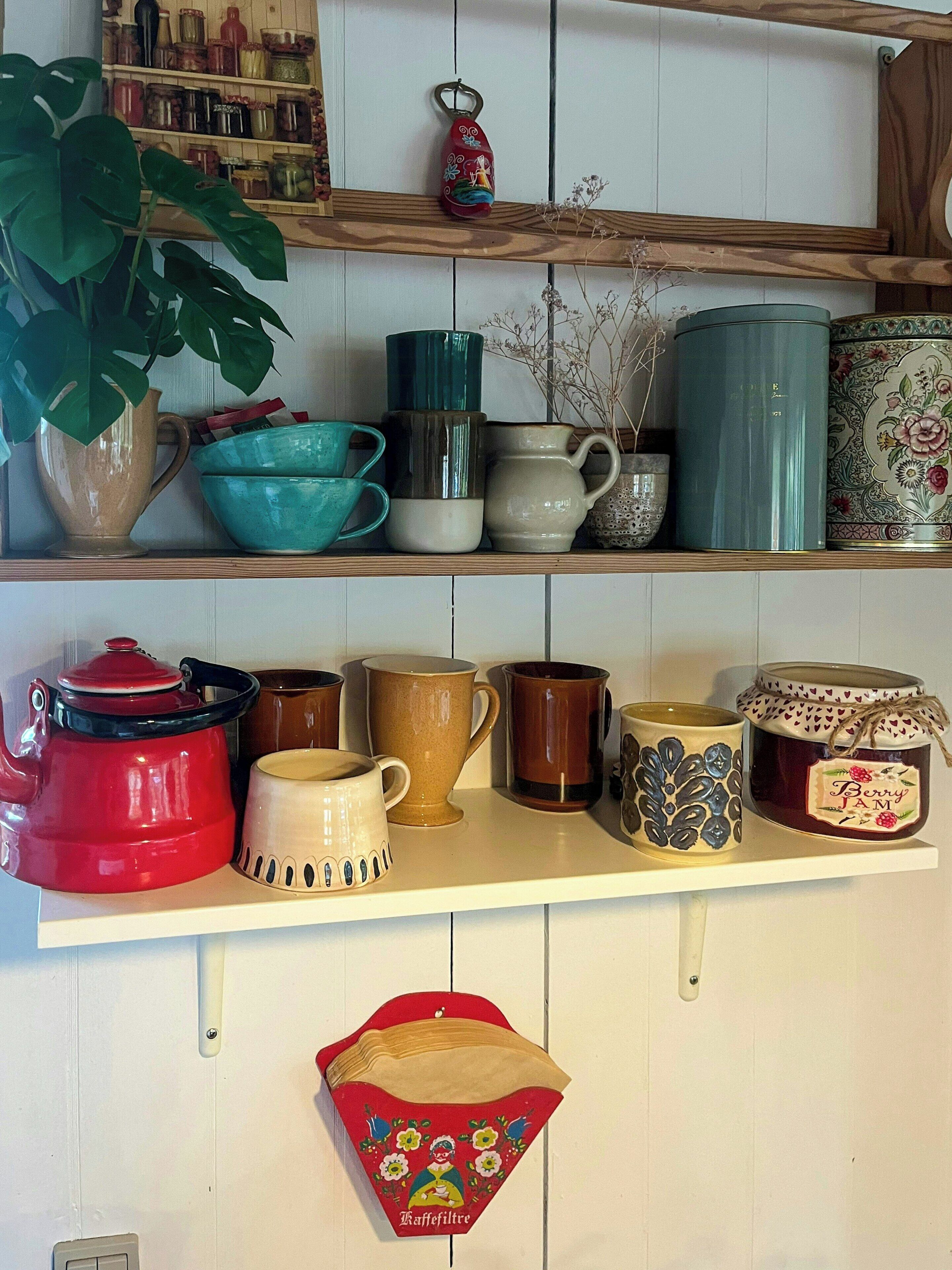 Un estante de cocina con diversos utensilios, incluyendo un hervidor rojo, un tarro de mermelada y varias tazas.