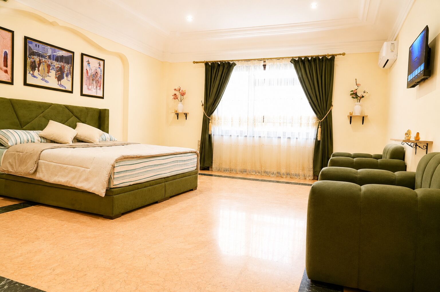 Habitación de hotel con cama, sillones verdes, televisor y obras de arte en la pared.