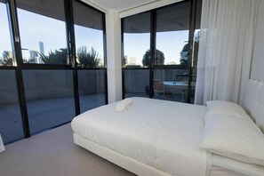 Ein Schlafzimmer mit einem großen Bett, Blick auf die Stadt durch raumhohe Fenster und einem Balkon mit Stuhl und Tisch.