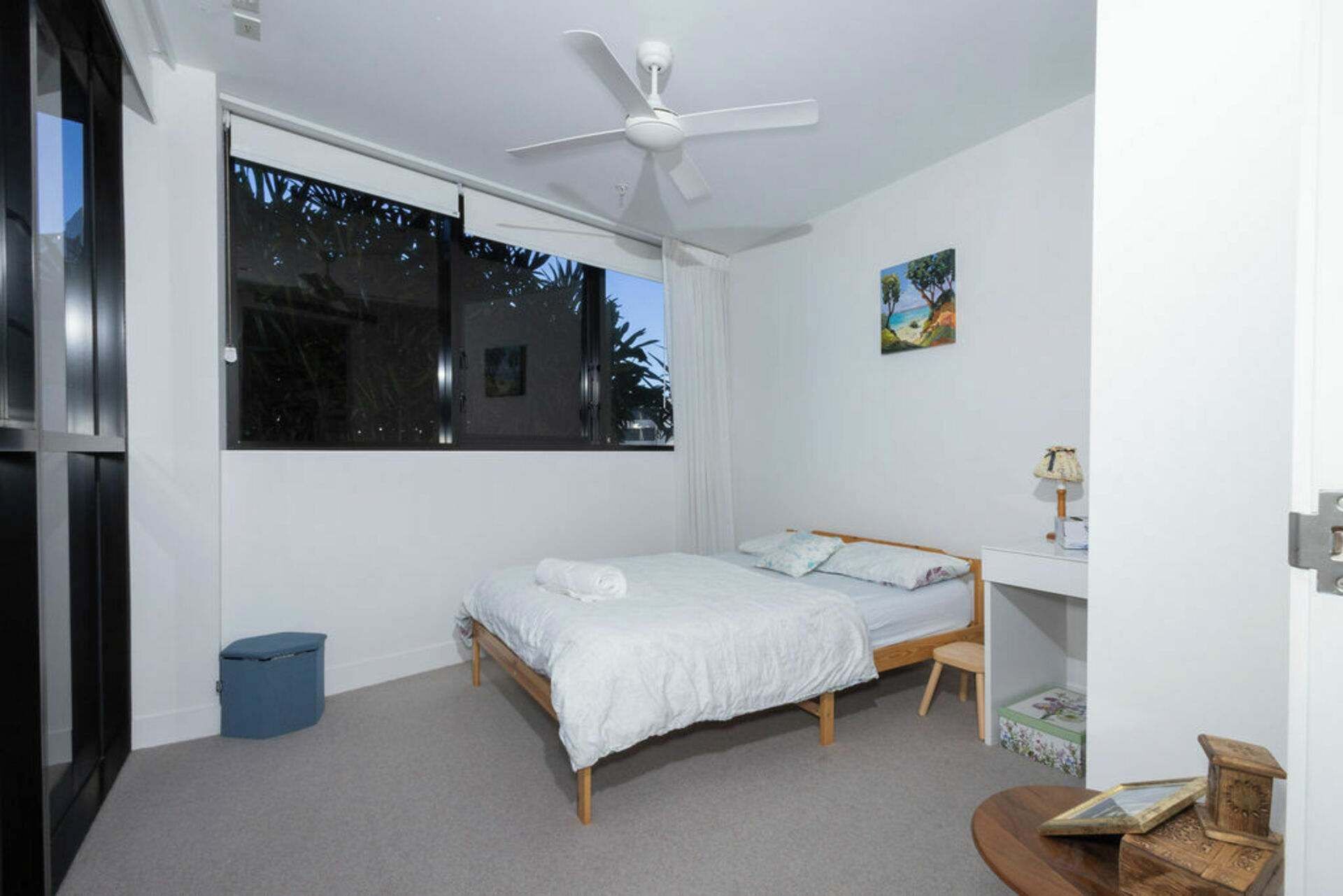 A bedroom with a bed, a desk, a chair, a ceiling fan, and a window with a view of trees.