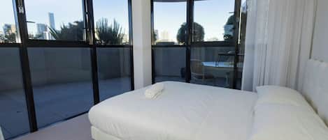 Une chambre d’hôtel dotée d’un grand lit, offrant une vue sur la ville grâce à de larges fenêtres du sol au plafond, et disposant d’un balcon avec une chaise et une table.