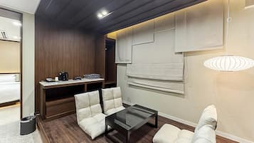 A modern hotel room with a wooden ceiling, a glass coffee table, two white cushioned chairs, a small desk with a coffee maker, and a large window with blinds.