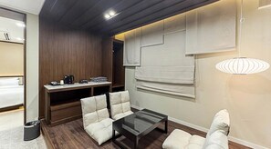 Une chambre d’hôtel moderne avec un plafond en bois, une table basse en verre, deux fauteuils blancs rembourrés, un petit bureau avec une machine à café et une grande fenêtre avec des stores.