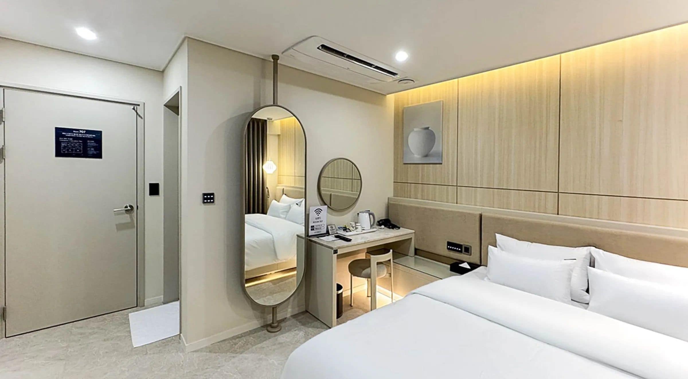 A hotel room with a bed, a desk with a chair, a mirror, a door, and a wall-mounted artwork.
