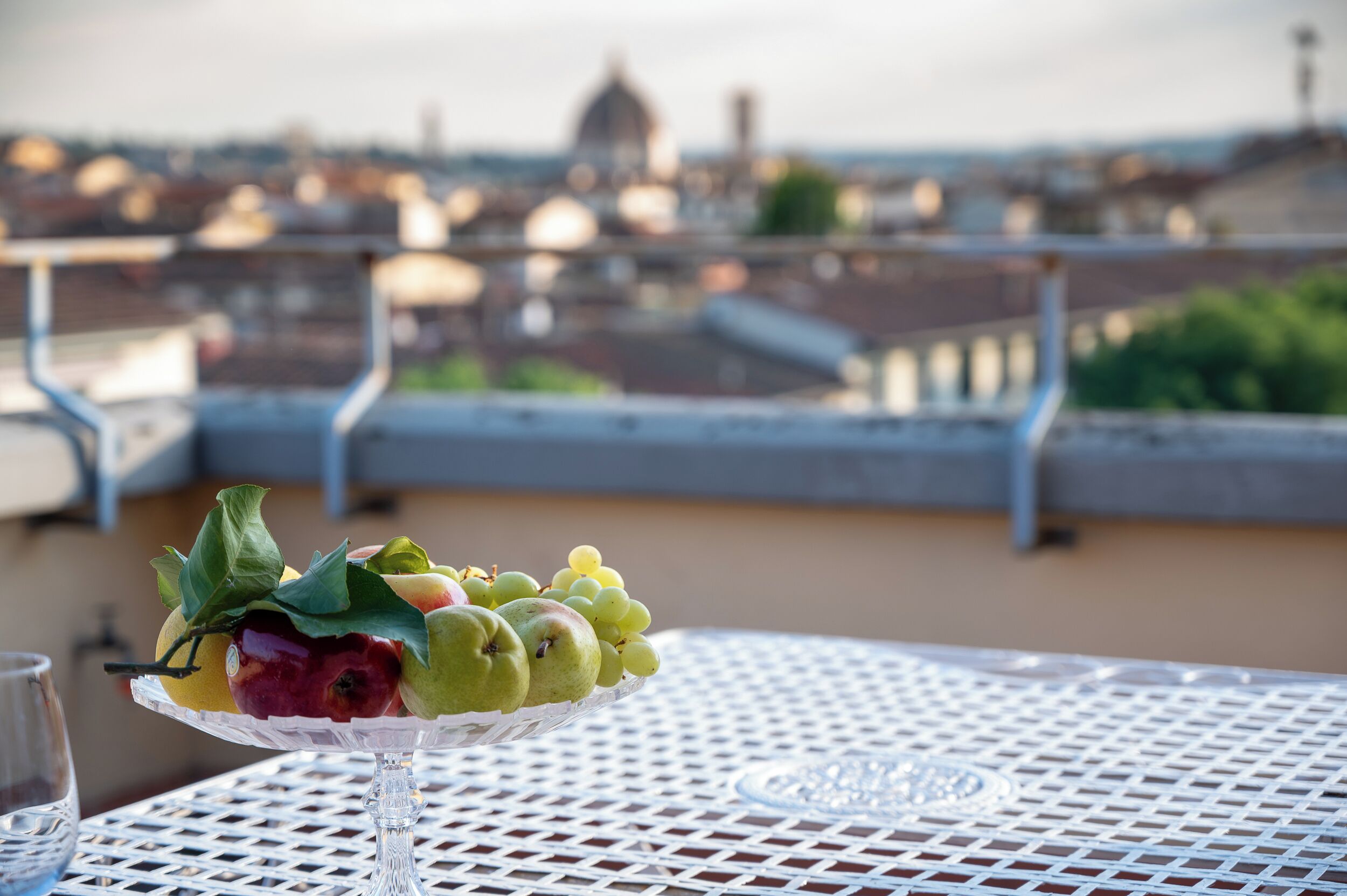A table with a fruit arrangement and a glass of water, overlooking a cityscape with buildings and a dome structure.