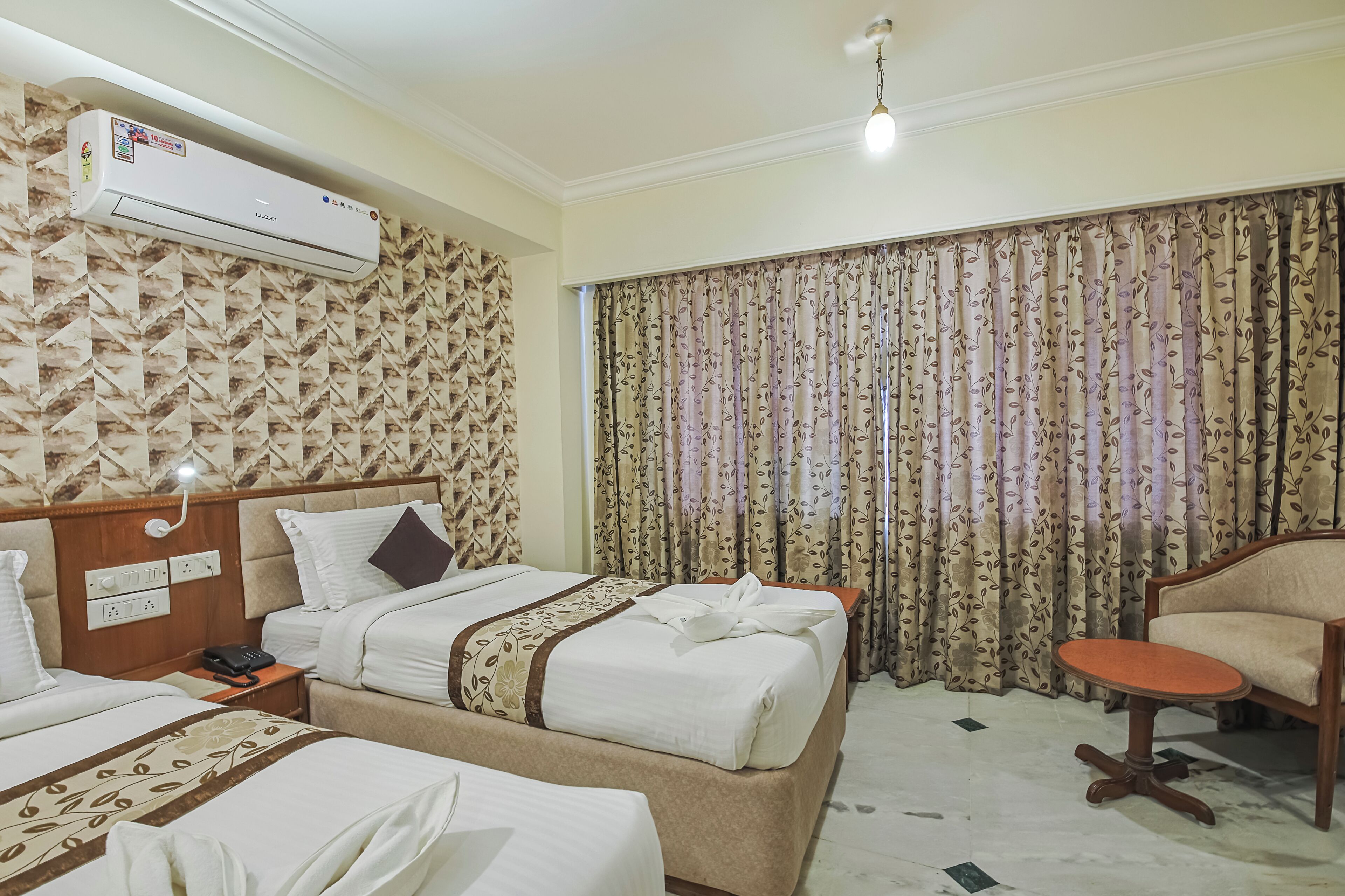 A hotel room with a bed, a bedside table with a phone, an air conditioning unit, a chair, and a window with curtains.