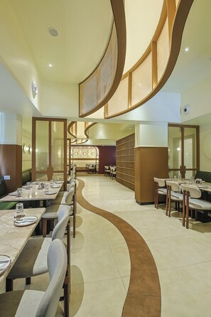 A modern restaurant with a curved ceiling design, multiple dining areas, and a central seating arrangement. - Hotel Maurya Residency (Mysore)