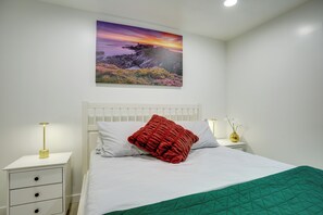 Ein Schlafzimmer mit einem weißen Bett, einem roten Kissen, einer grünen Steppdecke, einem weißen Nachttisch, einer goldenen Lampe und einem Wandbild.