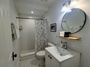 Ein Badezimmer mit Toilette, Dusche und Waschbecken. Über dem Waschbecken hängt ein Spiegel und darüber befindet sich ein Ablagefach mit einer Kosmetiktasche.