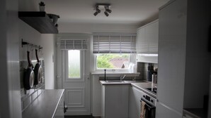 A compact kitchen with white cabinets, a built-in oven, and a window with blinds. - The Lomond House (Balloch)