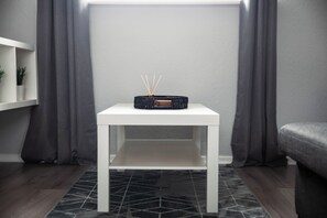 Uma mesa branca com um objeto preto em cima, uma parede cinzenta e cortinas cinzas.