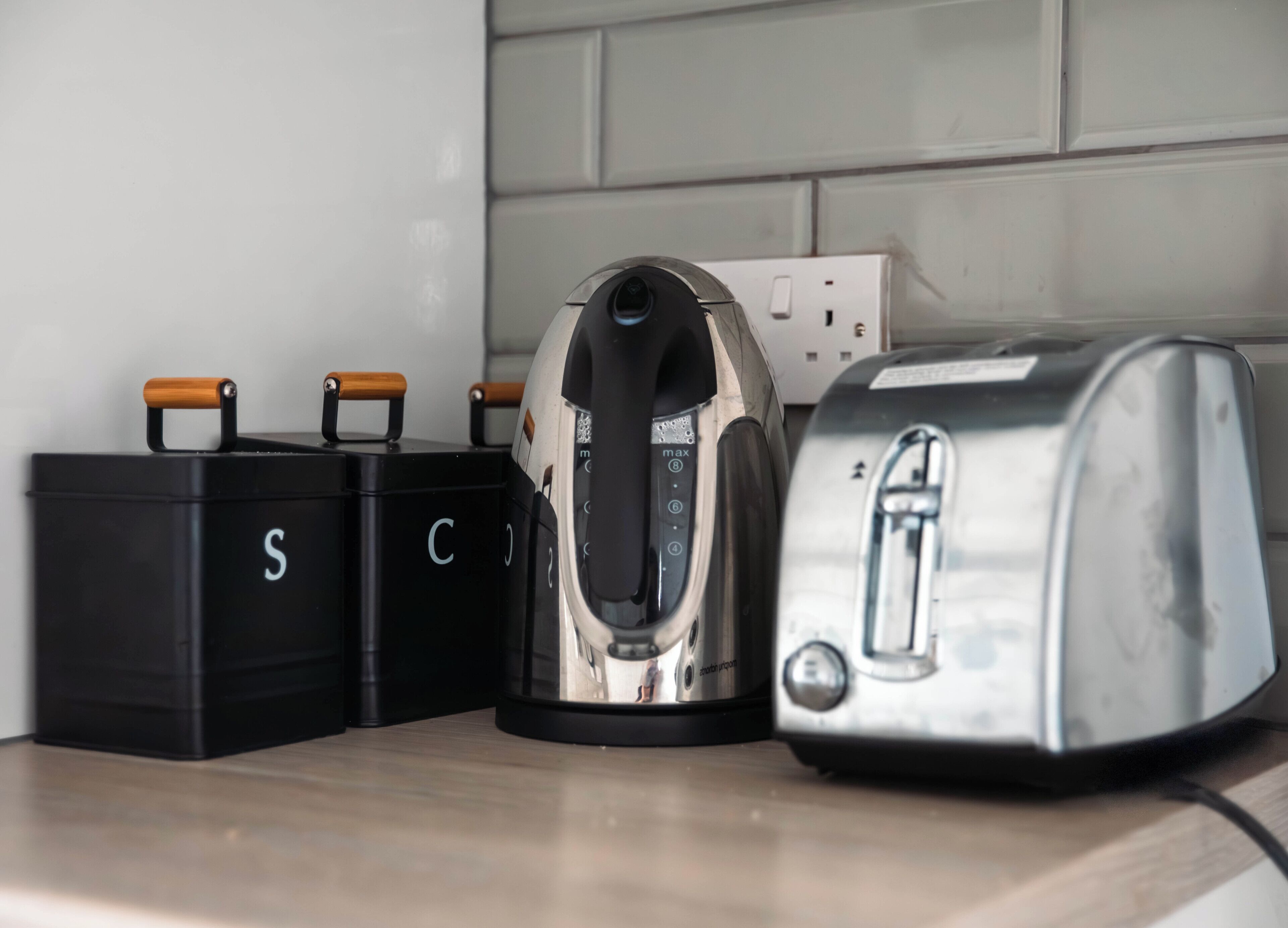 A kitchen counter with a kettle, a toaster, and two black storage containers with handles.