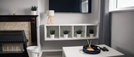 A living room with a fireplace, a television mounted on the wall, a white table with a tray, and a window with curtains.