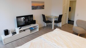 A hotel room with a bed, a TV, a desk with two chairs, a painting on the wall, and a door leading to another room. - Cozy one-room apartment right on the market square and next to the spa gardens (Grömitz)