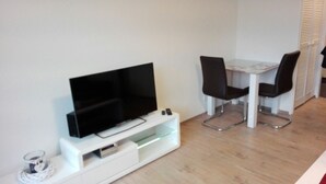A compact living space with a flat-screen TV on a white stand, a small table with two chairs, and a white cabinet with drawers. - Cozy one-room apartment right on the market square and next to the spa gardens (Grömitz)