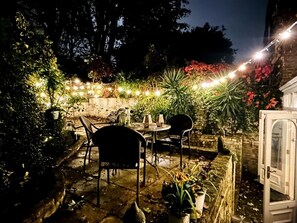 An outdoor patio area with stone flooring, metal chairs, a table, and string lights. - Kensington Garden Sanctuary - birds, squirrels & unique art! By Holland Park (Greater London)