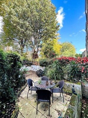 Eine Terrasse im Freien mit einem runden Tisch und vier Stühlen, umgeben von üppigem Grün und verschiedenen Pflanzen.