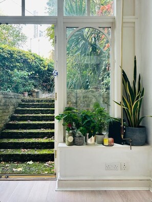A room with a large window overlooking a staircase leading to a garden. There are several potted plants on a windowsill. - Kensington Garden Sanctuary - birds, squirrels & unique art! By Holland Park (Greater London)
