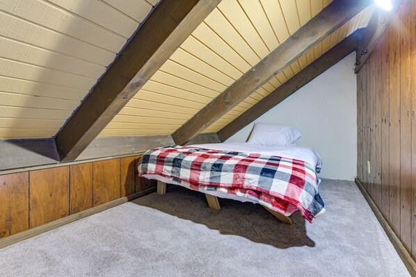 Ein kleiner, schräger Raum mit einem Einzelbett, einem hölzernen Kopfteil und einer beigen Decke.
