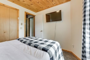 Un dormitorio con cama, techo de madera, televisor montado en la pared y una puerta.