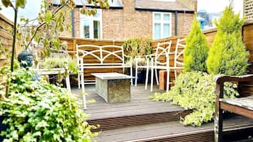 A wooden deck with a white metal pergola, a stone bench, and green plants.