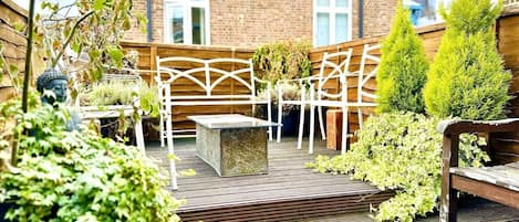 A wooden deck with a white metal pergola, a stone bench, and green plants.
