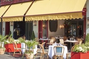 An outdoor cafe with tables and chairs, a yellow awning, and potted plants.
