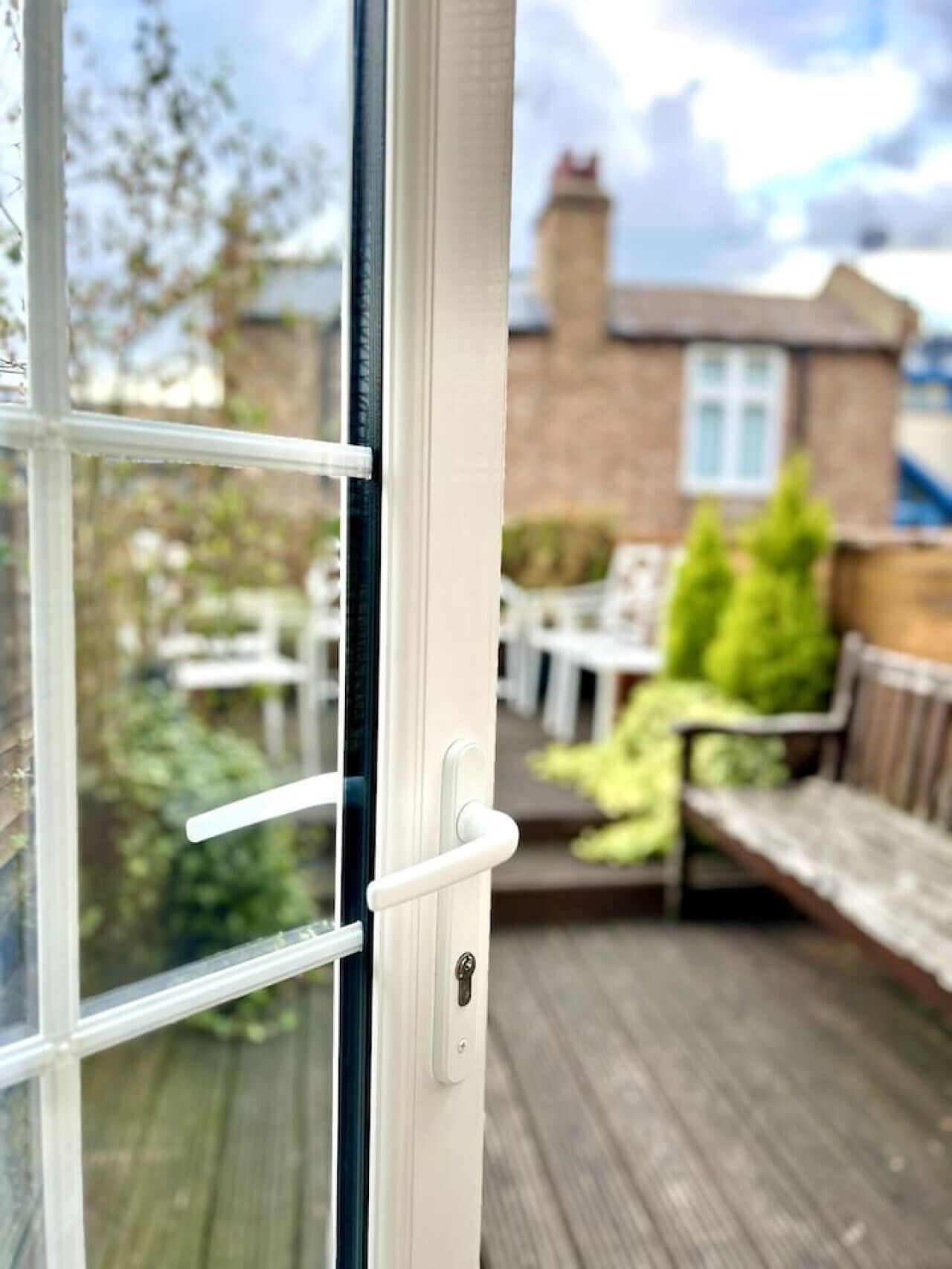 A view through a window with a white frame and a door handle, leading to an outdoor patio area with a wooden deck, a bench, and a table.