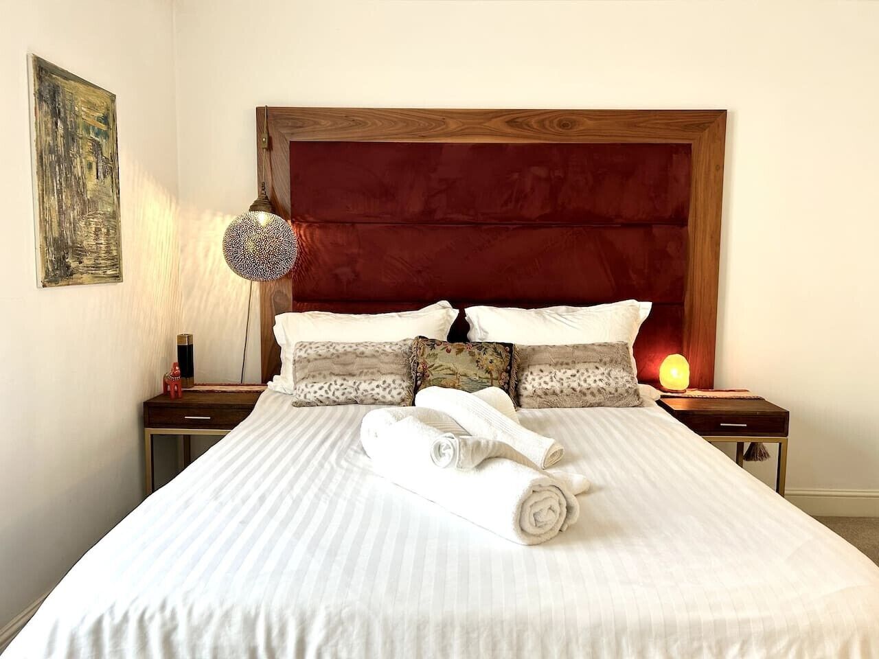 A neatly made bed with rolled towels, a wooden headboard, and a wall-mounted lamp.