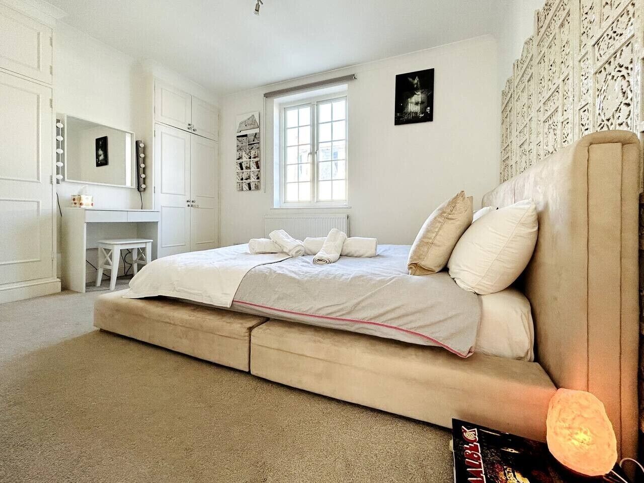 A bedroom with a bed, a vanity with a mirror, a window, and a wall with a decorative panel.